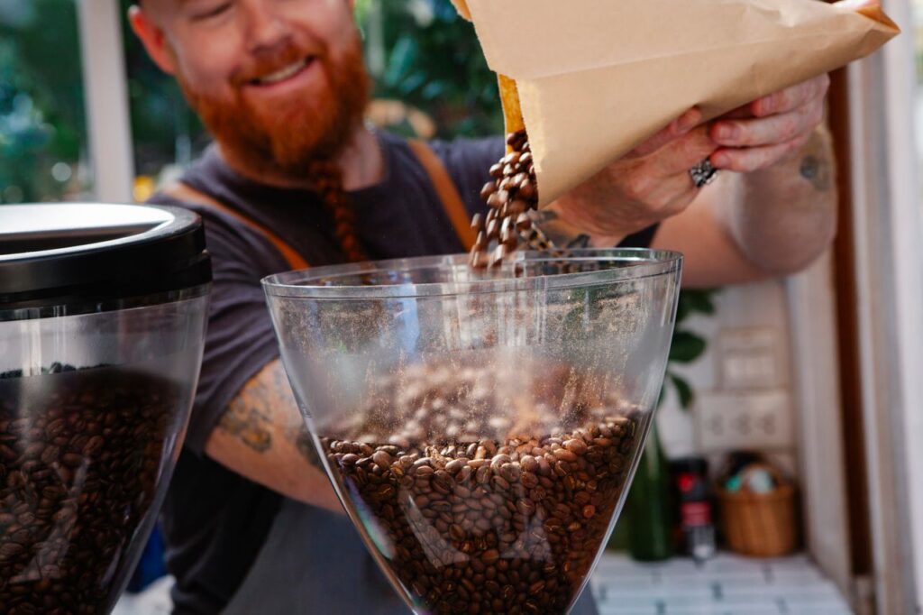 Crop smiling unshaven barista pouring roasted coffee beans into transparent grinder at work in cafeteria